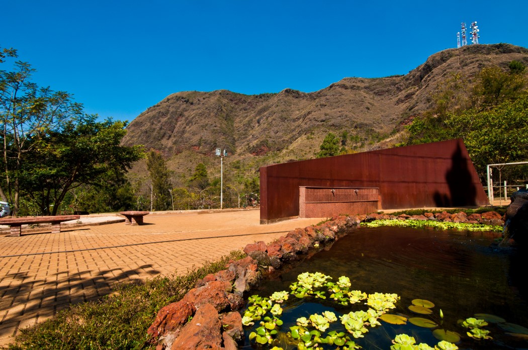 Área do Parque Serra do Curral. Em primeiro plano, um lago com plantas aquáticas e uma parede de metal enferrujado com chafarizes. Ao lado, um pátio de tijolos e a terra batida, com bancos de madeira. O local está na base de uma grande montanha, com a encosta em tons de marrom e verde. O céu está azul e sem nuvens.