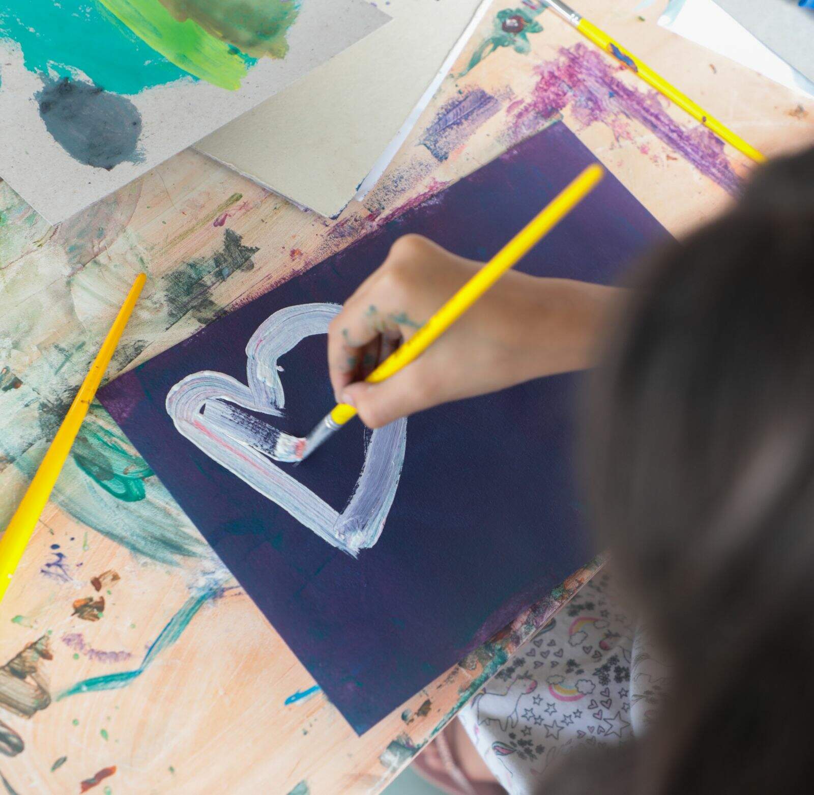 Imagem mostra uma criança de costas no canto da imagem pintando em um papel preto. O foco da fotografia esta principalmente na pintura da menina. É um coração branco em uma folha preta.