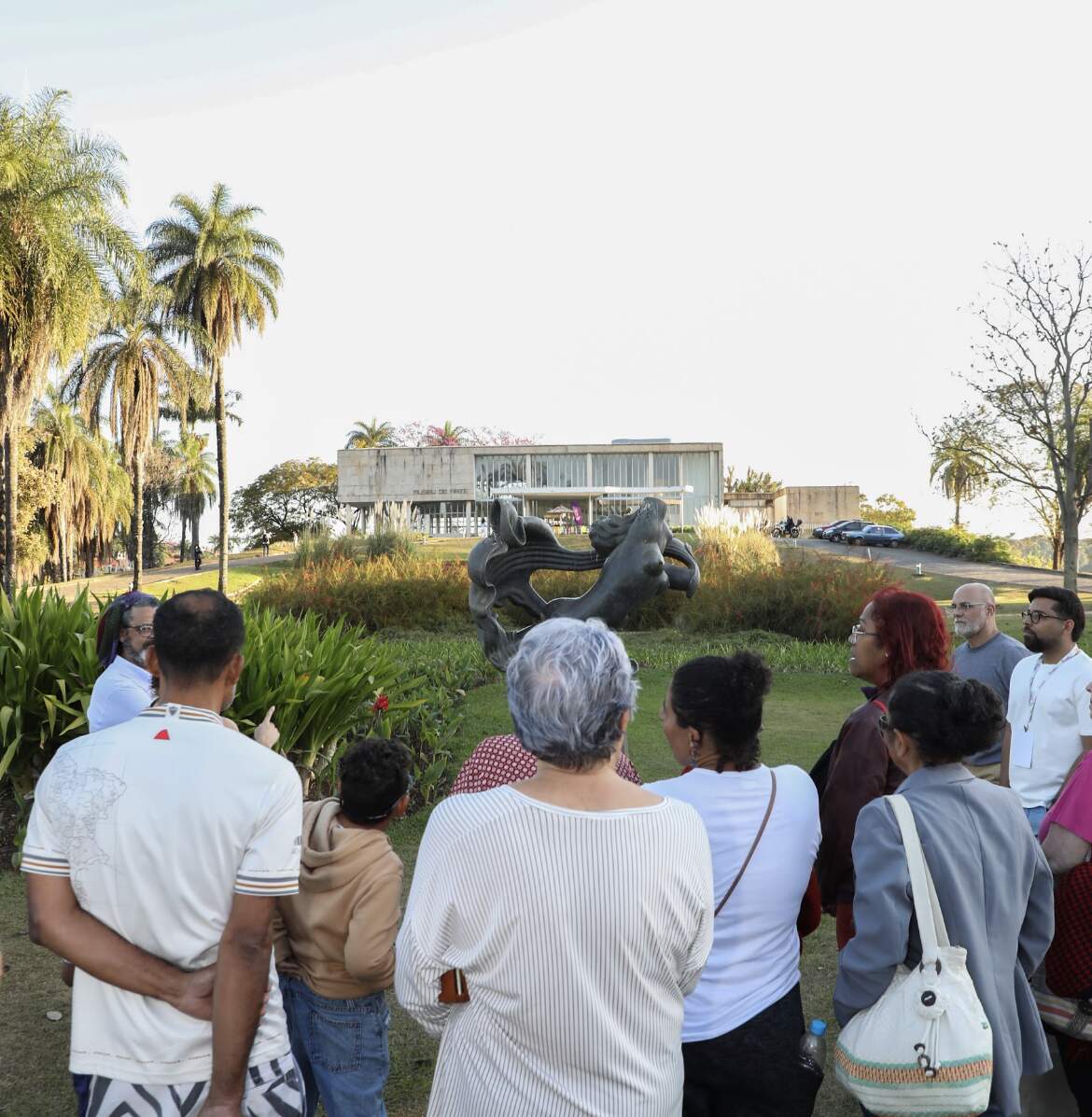 Grupo de pessoas admira os jardins do Museu de Arte da Pampulha. Elas estão de costas para a câmera olhando de frente ao Museu