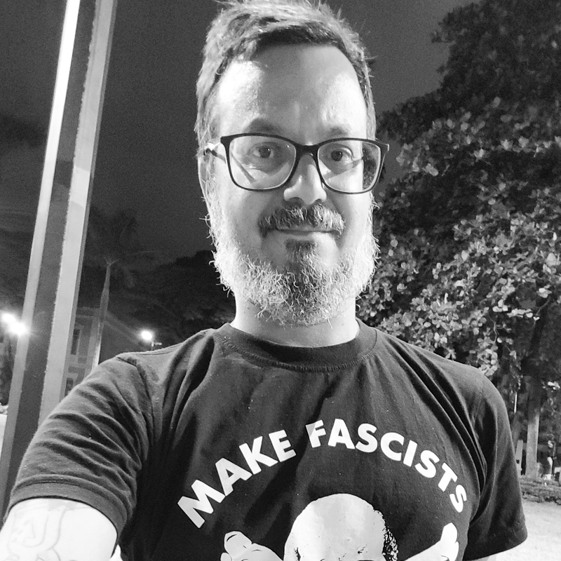 Uma selfie com um homem em pé, vestido de blusa preta com o desenho de uma caveira escrito make facists. Está à noite e ele está na rua. 