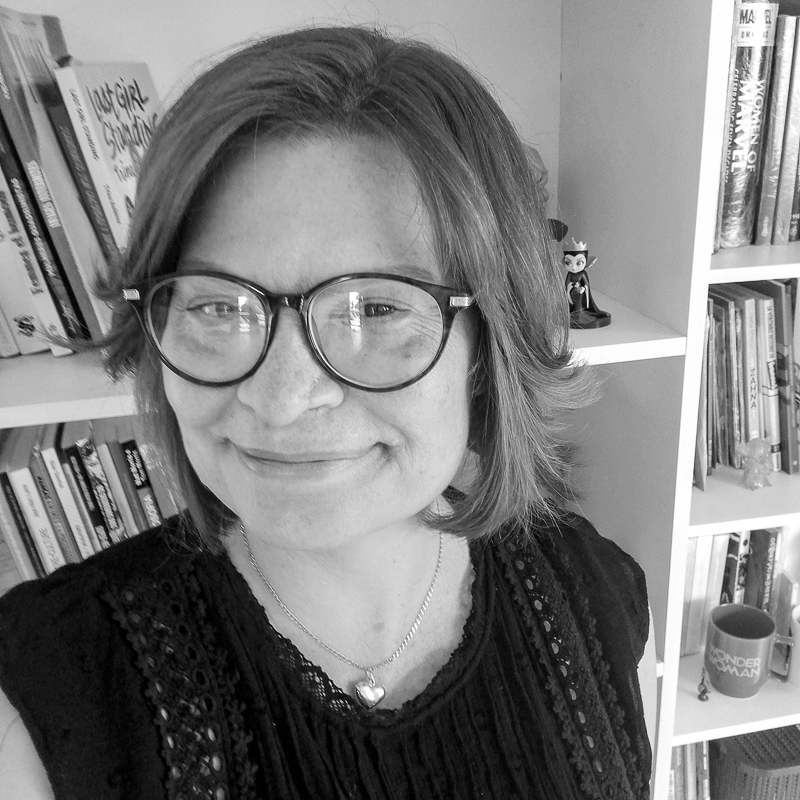 Selfie com uma mulher de blusa preta e óculos de grau, sorrindo em frente a uma estante de livros.