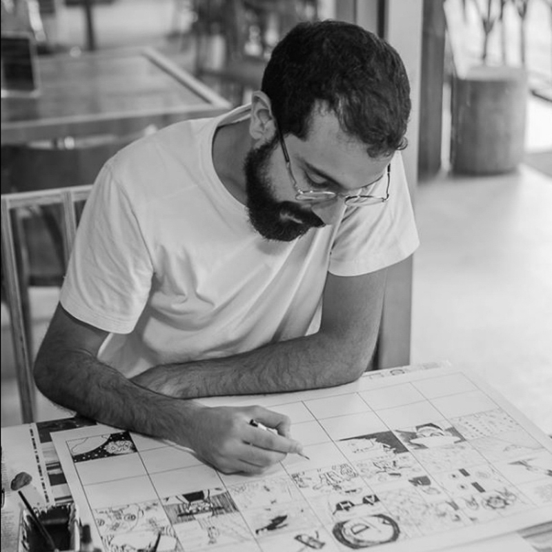 Fotografia preto e branca, com um homem sentado executando alguns desenhos de histórias em quadrinhos. Ele está concentrado e olhando para o desenho.