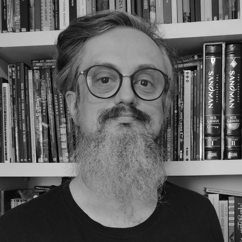 Homem com barba grande e óculos olhando para a câmera em frente a uma estante de livros