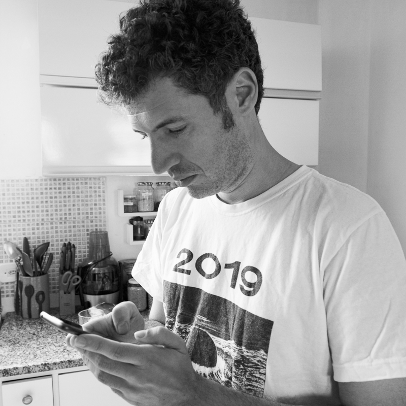  Homem em pé em uma cozinha, usando uma blusa branca e preta escrita 2019. Ele está olhando e mexendo no celular.