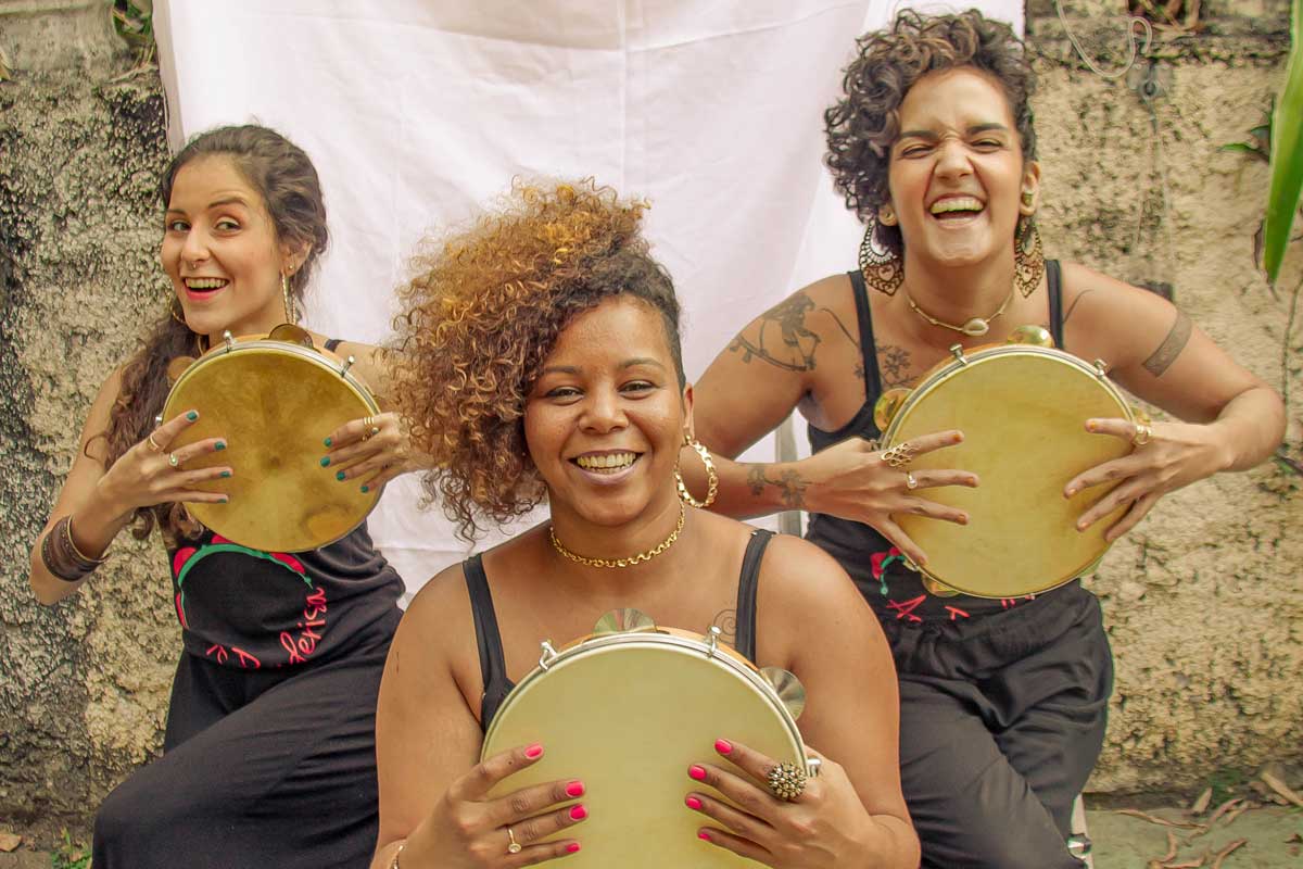 A foto mostra quatro integrantes do grupo tocando pandeiros durante uma apresentação em local aberto, é possível identificar o rosto de duas delas, a da direita está sorrindo, possui cabelos lisos brancos curtos e usa óculos de grau. A da esquerda tem cabelos crespos, castanhos e usa batom vermelho.