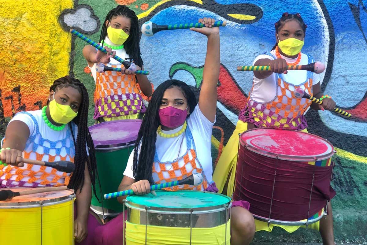  A foto mostra quatro mulheres, em frente a uma parede grafitada, segurando tambores. As duas que aparecem à frente estão agachadas, com os braços erguidos. As que aparecem atrás estão em pé, com os braços erguidos apontando para câmera. Todas seguram baquetas de tambor e usam máscaras de proteção 