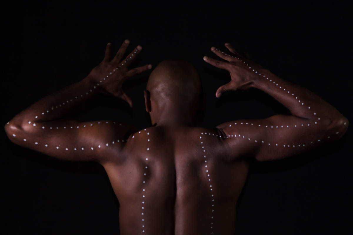 A foto é preto e branco e mostra o artista, que é um homem negro, careca. Ele está em pé, dentro de uma bacia de barro, num fundo preto. Ele está olhando para a câmera, usa sunga preta, está com o tronco curvado para frente e os braços esticados para trás 