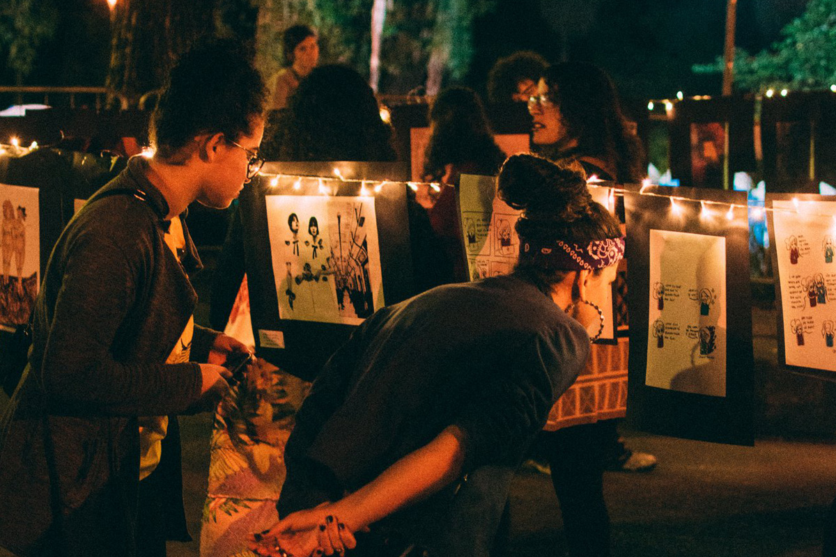 A foto mostra uma exposição de arte na rua. Há varais iluminados onde as obras, que têm fundo preto, estão penduradas. No primeiro plano da imagem, duas mulheres observam algumas obras. Ao fundo, outras pessoas também admiram as obras.