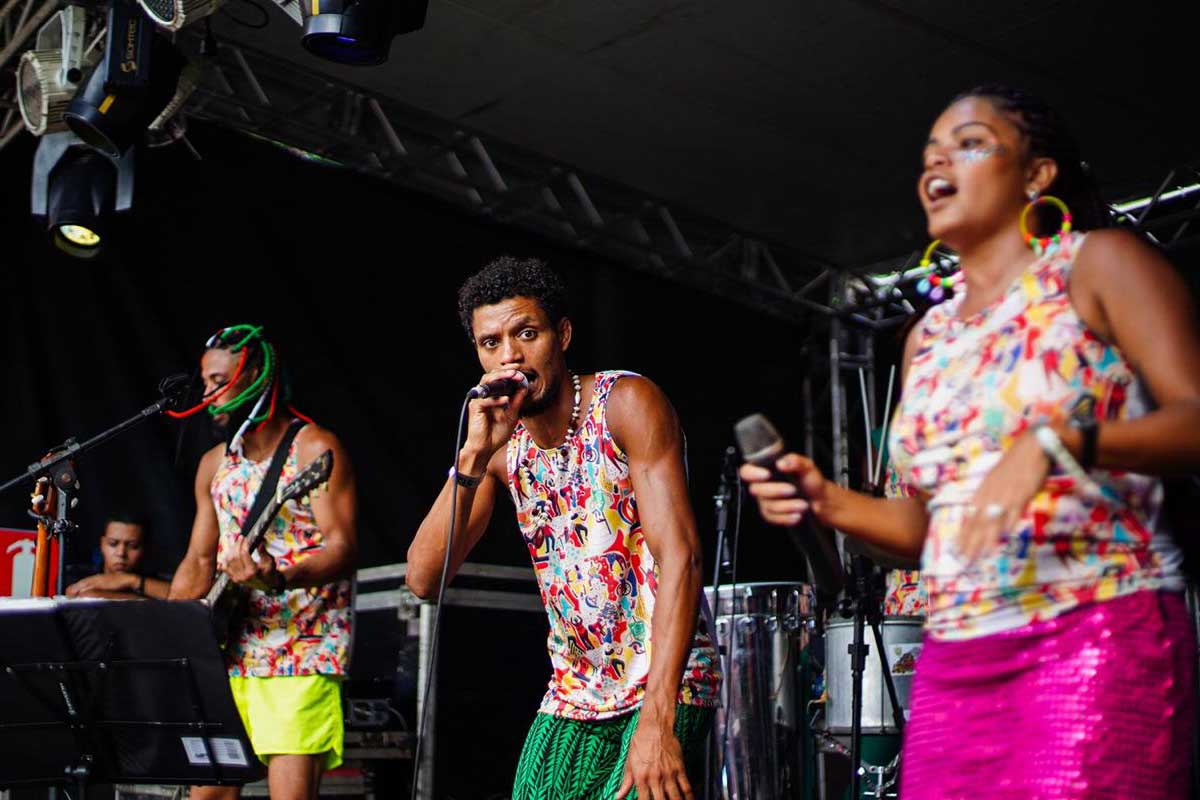 A foto mostra dois homens e uma mulher no palco, com microfones e camisas iguais. À esquerda, o homem tem dreads pretos, brancos, verdes e vermelhos, usa short amarelo e toca baixo. Ao meio, o homem usa short verde e colar e canta. À direita, a mulher usa saia rosa, brincos grandes e brilho na face.