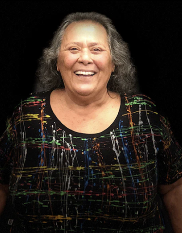 Foto de uma mulher de cabelos grisalhos na altura dos ombros e sorriso largo posando para foto. A mulher veste uma blusa preta com esguichos finos de tinta colorida. O fundo da foto é preto e a mulher está em pé e aparece da cintura para cima.