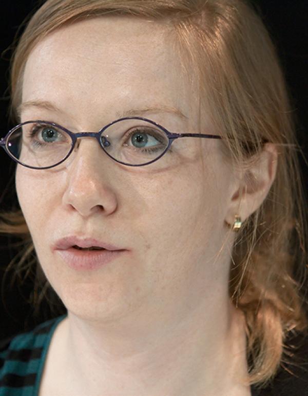 Foto do rosto de uma mulher. A mulher tem cabelos lisos e loiros, olhos azuis e usa óculos de grau com armação roxa, fina e em forma amendoada. A mulher usa pequenos brincos de argola dourada. A mulher está olhando ao longe, com a boca entreaberta, como se estivesse no meio de uma fala.