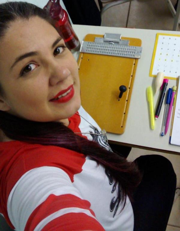 Foto de uma mulher sorrindo posando para uma selfie. A mulher tem cabelos pretos longos e lisos amarrados em um rabo de cavalo lateral. A mulher usa batom vermelho e blusa branca com mangas com detalhes vermelhas.