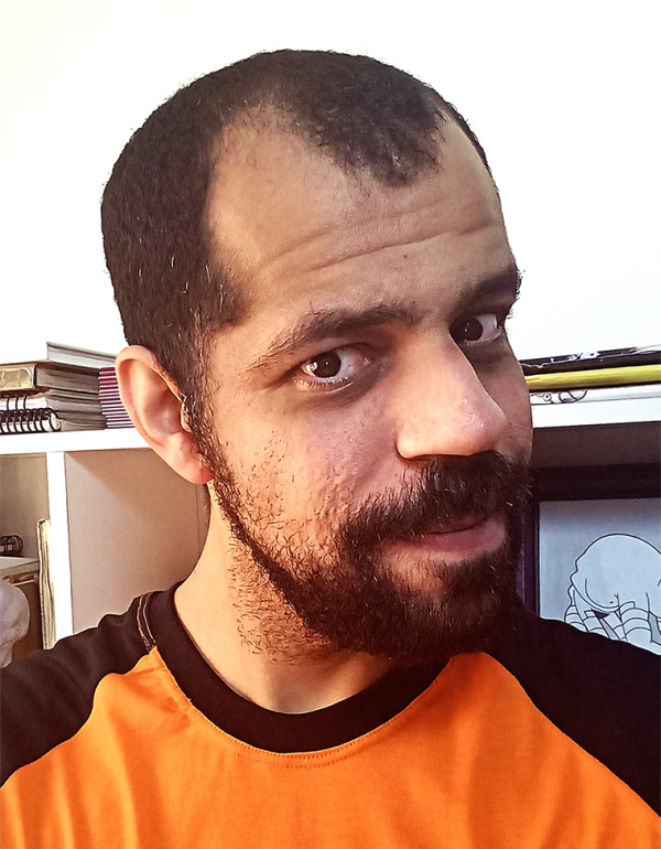 Foto do rosto de um homem. O homem tem barba e cabelos pretos, curtos e com entradas. O homem veste blusa laranja com mangas e gola azuis escuras. O homem posa para a foto sem sorrir. Ao fundo uma parede branca e uma estante com livros deitados. 