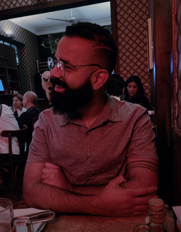 Foto de um homem sentado, com os braços cruzados sobre uma mesa, sorrindo e olhando para o lado. O homem tem cabelos curtos pretos, raspados do lado, e barba preta grande. O homem veste blusa marrom calara, social de mangas curtas, com bolinhas brancas. Atrás do homem tem pessoas sentadas em mesas, num ambiente que parece um restaurante.