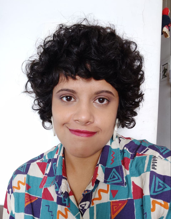 foto do rosto de uma mulher branca, de cabelos castanhos, acima dos ombros, ondulados, com franja. A mulher veste camisa colorida de gola com estampa geométrica em tons de azul e vermelho. A mulher usa brincos brancos e batom rosa. A mulher posa para foto sorrindo sem mostrar os dentes. Atrás dela uma parede branca.