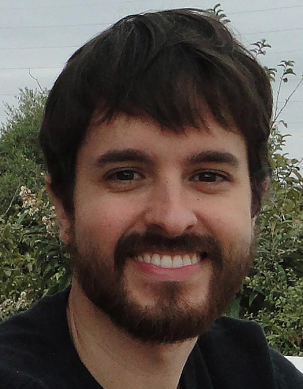 Descrição da imagem: foto do rosto de um homem sorrindo. O homem tem cabelos castanhos lisos e curtos, com franja. O homem tem barba castanha e sobrancelhas grossas. O homem posa para foto com um sorriso no rosto. O homem veste um moletom preto. O homem está ao ar livre em frente a um fundo com várias folhagens. 