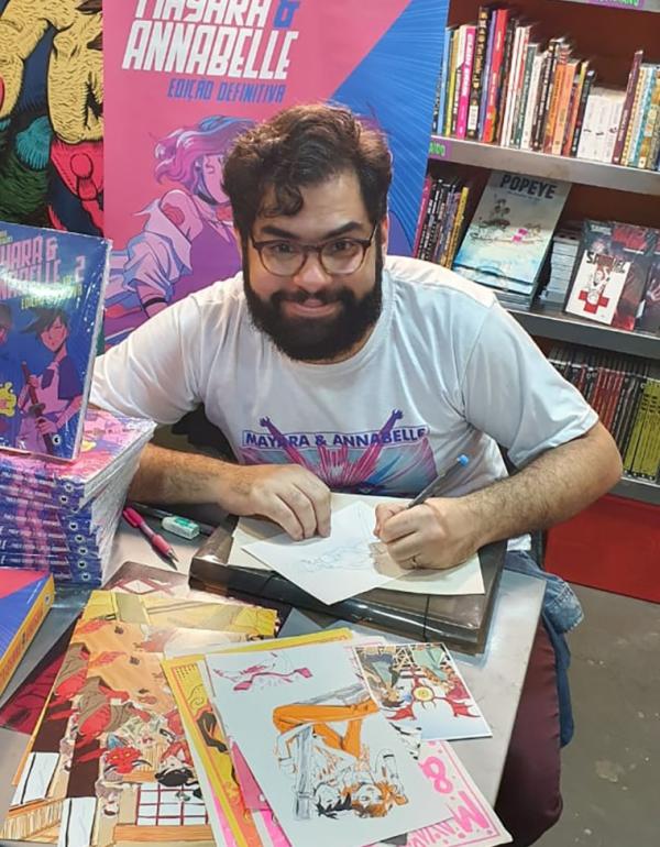 foto de um homem desenhando e sorrindo posando para a foto. O homem está sentado com as mão sobre uma mesa, desenhando num papel branco. O homem tem cabelos pretos lisos, sobrancelhas pretas grossas e barba preta cheia. O homem usa óculos de grau com armação redonda marrom. O homem sorri sem mostrar os dentes. O homem veste uma blusa branca de mangas curtas com uma ilustração na frente e o texto estampado: Mayara e Anabelle. 