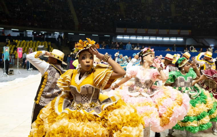 Um grupo de quadrilha junina se apresentando em um tablado, com destaque para uma mulher de vestido amarelo a frente da foto.