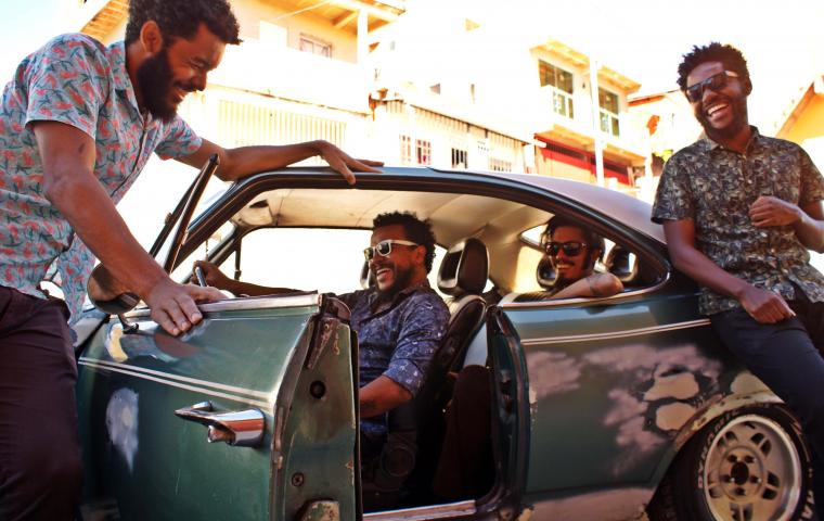 Foto de divulgação do grupo musical Diplomattas. Quatro homens vestem camisas de botão floridas, sorridentes, barbados, três deles usam óculos escuros. Dois deles estão dentro de um carro verde antigo. Os outros dois estão do lado de fora do carro, um segurando a porta aberta do carro, ao lado do motorista e outro está encostado do lado de fora.