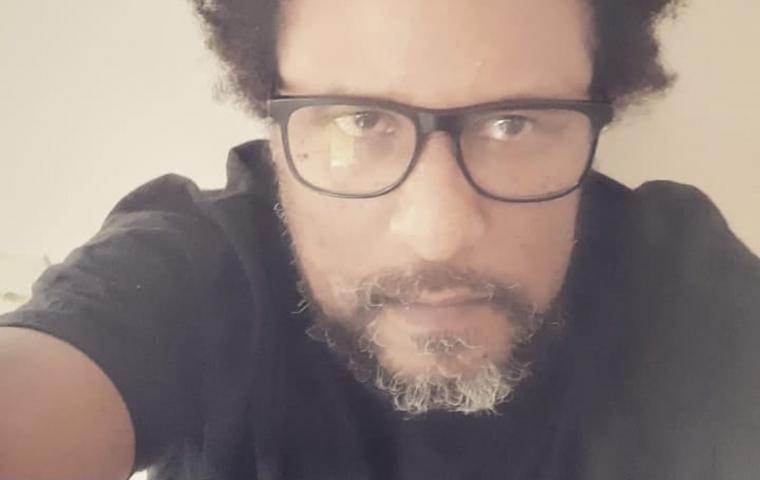 Selfie de homem negro, de cabelos pretos crespos, barba levemente grisalha. O homem usa óculos de grau com armação preta e veste blusa de mangas curtas pretas.