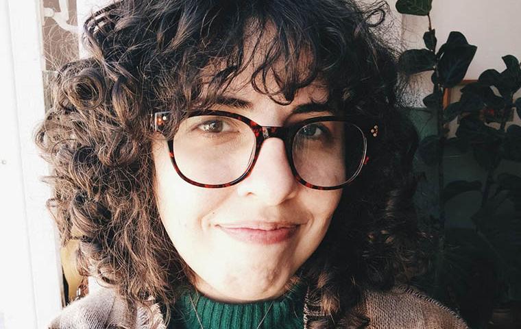 Descrição da imagem: foto do rosto de uma mulher branca, de cabelos castanhos cacheados, acima dos ombros, com franja. A mulher usa óculos de grau com armação redonda marrom escura com manchas pretas. A mulher veste casaco de lã em tons de marrom com blusa verde canelada com gola alta por dentro. A mulher posa para foto sorrindo sem mostrar os dentes. Atrás dela uma parede branca e uma planta no seu canto esquerdo.