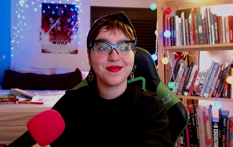 foto de uma mulher dos ombros para cima. A mulher tem cabelos castanhos claros e curtos e sobrancelhas grossas escuras. A mulher usa lenço preto na cabeça, em forma de arco, óculos de grau com armação grossa preta e batom vermelho. A mulher usa blusa de gola alta preta e brincos de argola acobreados. A mulher está sentada numa cadeira gamer preta e verde. Ao fundo, um ambiente desfocado, é possível ver atrás da mulher uma janela fechada preta, uma prateleira e várias luzinhas estilo luzinha de natal. 