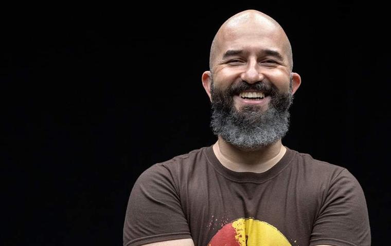 Foto de um homem sorridente, aparecendo dos cotovelos para cima, com os braços cruzados. O homem é careca, tem sobrancelhas grossas castanhas, bigode e barba grande levemente grisalha. O homem sorri largamente, com os olhos espremidos. O homem veste blusa marrom, com estampa amarela com detalhe vermelho na frente. O fundo da foto é preto. 