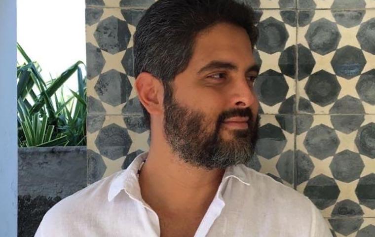 Foto de um homem de barba e cabelos pretos curtos, levemente grisalhos, sobrancelhas grossas pretas, olhando para o lado, vestindo blusa social branca de mangas curtas e colarinho desabotoado. Atrás do homem uma parede com pintura geométrica nas cores branco e cinza.