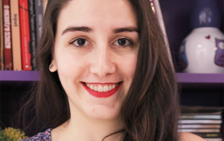  foto de uma mulher aparecendo dos ombros para cima. A mulher tem cabelos castanhos claros lisos e longos. A mulher usa batom vermelho, um piercing de argola no nariz e uma regata azul-escura de estampa florida. A mulher posa para a foto sorrindo em frente a uma estante cheia de livros. 