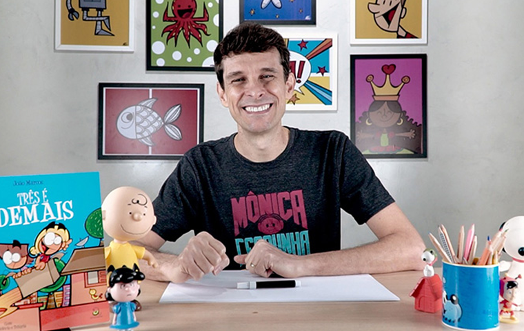 Descrição da imagem: foto de um homem branco com sorriso largo posando para a foto. O homem tem cabelos castanhos curtos e lisos. O homem está sentado e veste uma blusa preta de mangas curtas, com estampa do nome e do rosto da personagem Mônica do Maurício de Souza, na cor vermelha. Abaixo do rosto da Mônica está escrito uma palavra em azul que não é possível identificar. O homem está com os braços sobre uma mesa de madeira, com um papel branco em cima dela. No canto esquerdo da mesa um boneco do Charlie Br