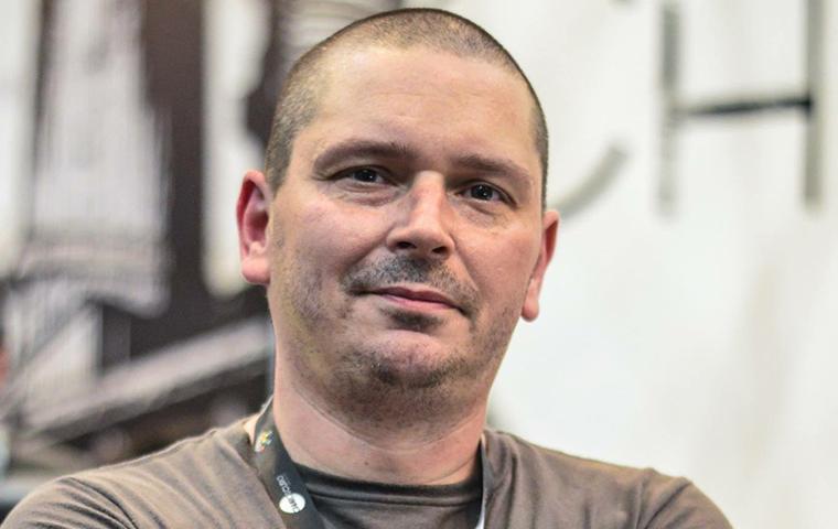 Foto de um homem posando para foto de braços cruzados, esboçando um sorriso de boca fechada. O homem é branco careca e sem barba. O homem usa blusa cinza de mangas curtas, relógio marrom no pulso direito e tem nos braços tatuagens na cor preta. 