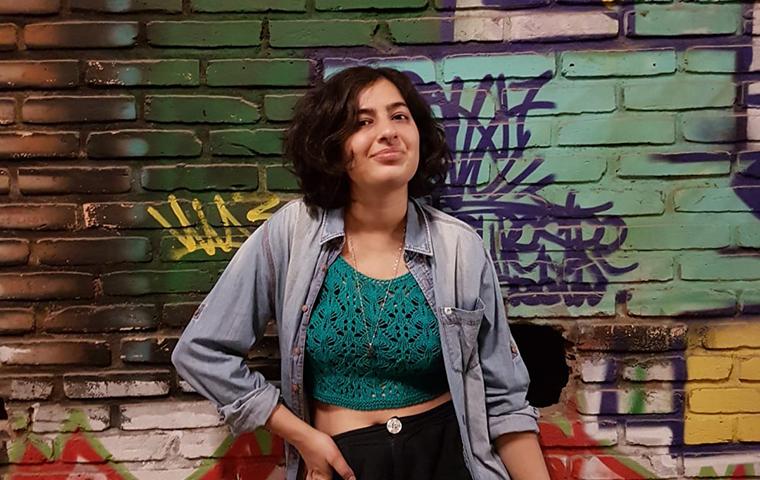 foto de uma mulher em pé, em frente a um muro grafitado. A mulher tem cabelos pretos ondulados, curtos, em corte chanel abaixo das orelhas. A mulher posa para foto esboçando um sorriso. A mulher veste uma camisa jeans de mangas compridas aberta, com uma regata verde de renda por baixo e barriga de fora. A mulher veste uma calça de tecido preta e usa um colar longo dourado com um pequeno pingente triangular. Ao fundo, um mundo grafitado colorido com alguns escritos indecifráveis. 