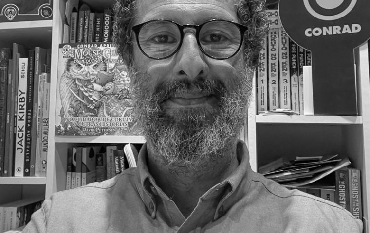  Selfie de um homem de barba em frente a uma estante de livros.
