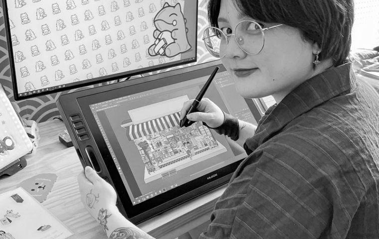 mulher jovem de cabelo liso e traços orientais está sentada de lado olhando para a câmera. Ela está em frente ao computador e outros equipamentos criando um desenho.