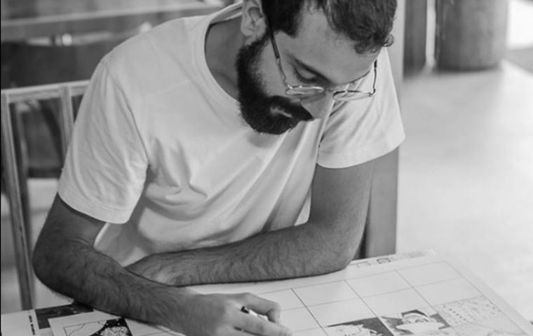 Fotografia preto e branca, com um homem sentado executando alguns desenhos de histórias em quadrinhos. Ele está concentrado e olhando para o desenho.