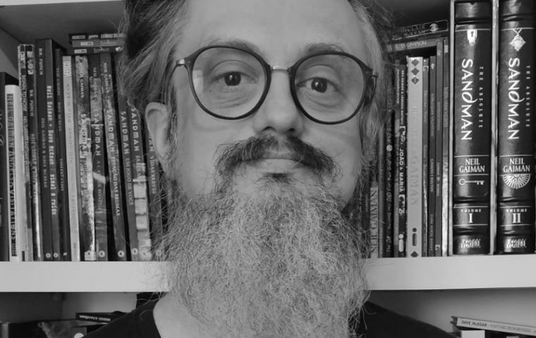 Homem com barba grande e óculos olhando para a câmera em frente a uma estante de livros
