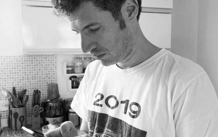  Homem em pé em uma cozinha, usando uma blusa branca e preta escrita 2019. Ele está olhando e mexendo no celular.
