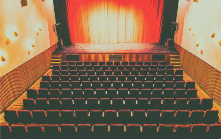 Teatro Marília com as luzes acesas. No enquadramento é possível ver o palco com cortinas fechadas e as cadeiras da platéia