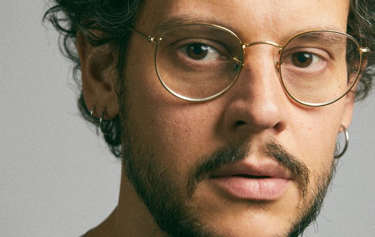 Fotografia de Vinícius de Souza, um homem branco, jovem, usa óculos de aros redondos e finos, bigodes, barba e cabelo castanhos..