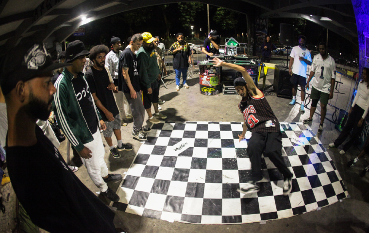 A imagem mostra um grupo de pessoas em um ambiente urbano, participando de uma competição ou batalha de dança em estilo hip-hop. No centro, um dançarino está realizando um movimento dinâmico sobre um tablado quadriculado, enquanto os outros participantes, em pé ao redor, assistem com interesse. O local parece iluminado por luzes artificiais, com uma mesa de DJ ao fundo e um clima animado de confraternização e arte de rua.