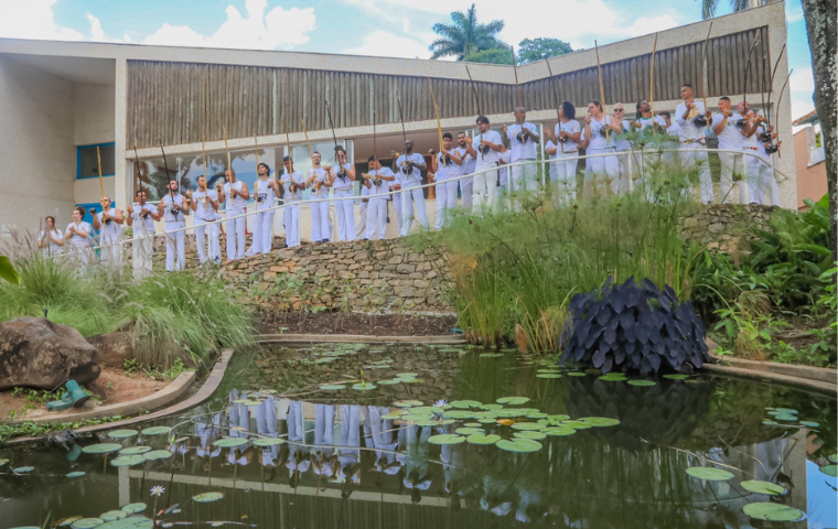 Na imagem um grupo de capoeira está vestido de branco tocando berimbau em frente à varanda do museu casa kubitschek. a foto enquadra em primeiro plano o lago do museu e em segundo plano os integrantes do grupo oficina de capoeira em linha reta tendo ao fundo o telhado borboleta da edificação.