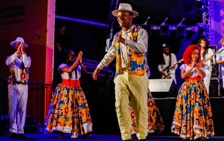 A imagem mostra um grupo de pessoas em um palco durante uma apresentação cultural vibrante e alegre. Um homem ao centro está sorrindo e dançando, vestindo um colete colorido estampado com girassóis e um chapéu branco, combinando com calças claras. Ele está cercado por outras pessoas também trajadas com roupas tradicionais. Ao fundo, músicos tocam ao vivo, dando energia à cena. O clima é de celebração e representa uma expressão rica da cultura popular brasileira.
