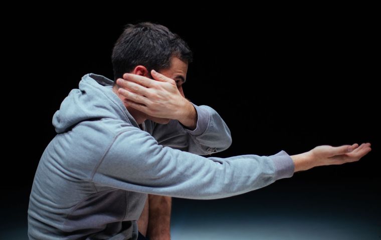 A imagem mostra o ator de moletom cinza em um ambiente escuro. Ele está de lado, com o braço direito estendido para frente, com a palma da mão voltada para cima. Sua mão esquerda cobre parcialmente seu rosto, escondendo um dos olhos e parte da bochecha. O fundo preto destaca ainda mais a figura e a expressividade do corpo e das mãos. A iluminação suave realça os contornos do rosto e das roupas, dando um efeito dramático à cena.