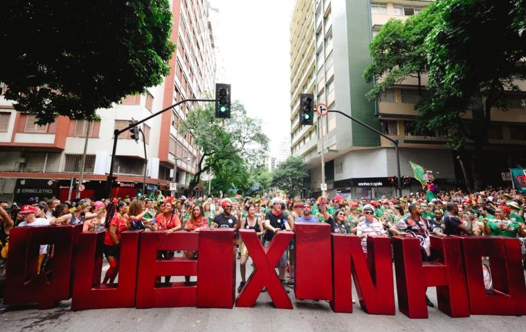A imagem mostra o bloco de carnaval desfilando por uma rua arborizada, cercada por prédios residenciais, em Belo Horizonte. A imagem mostra um grupo de pessoas segura grandes letras vermelhas tridimensionais que formam a palavra "QUEIXINHO". Também mostra uma multidão animada, vestindo roupas coloridas, adereços carnavalescos e fantasias. O ambiente é vibrante, com muitas pessoas usando acessórios como coroas de flores, óculos escuros e chapéus. A cena transmite alegria, diversidade e a energia contagiante.