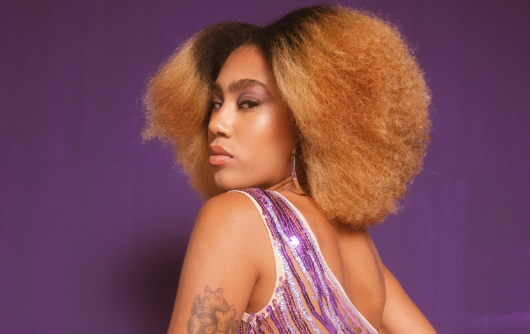 A foto mostra uma mulher negra e cabelos volumosos em um tom degradê de castanho escuro para loiro. Ela veste um vestido brilhante em tons de lilás e rosa, com um corte elegante que deixa um dos ombros à mostra. Seu olhar é marcante, voltado para a câmera numa expressão confiante e intensa. A maquiagem destaca os olhos com sombras em tons de roxo e um brilho. No braço, há uma tatuagem visível. O fundo da imagem é roxo, harmonizando com a paleta de cores do look, criando uma composição vibrante e sofisticada