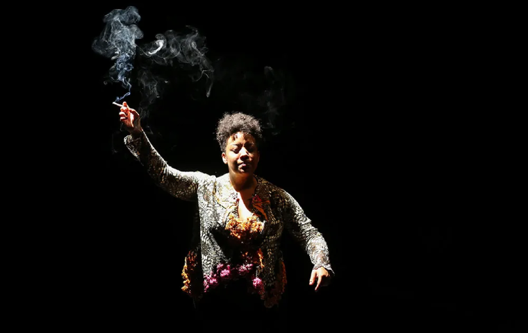 A imagem mostra uma pessoa no palco, iluminada dramaticamente contra um fundo escuro. Ela segura um cigarro aceso, com a fumaça subindo ao seu redor. Sua expressão e postura sugerem intensidade e emoção. A roupa tem detalhes brilhantes e elementos florais, adicionando textura e profundidade à cena. A iluminação focada realça sua presença e a atmosfera teatral da fotografia.