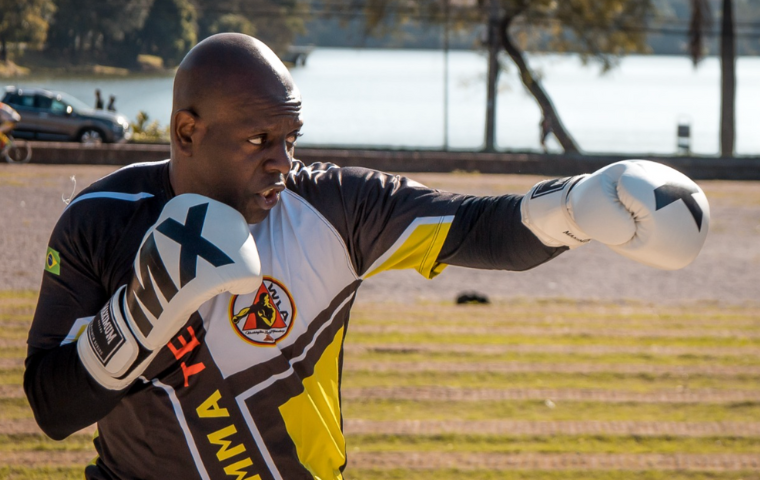 Na imagem um homem negro, careca, usa luvas brancas de boxe e roupas esportivas pretas com estampas amarela e branca. Ao fundo é possível ver a lagoa da Pampulha. Ele faz um movimento de boxe com uma das luvas próxima do rosto (DIREITA) e outra desferindo um golpe de soco no ar (ESQUERDA).)