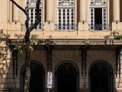 Dito pelo ChatGPT: Fachada de edifício antigo bege com colunas e janelas altas em arco. Na parte inferior, há três portas também em arco, uma placa de trânsito e uma árvore em frente à calçada.