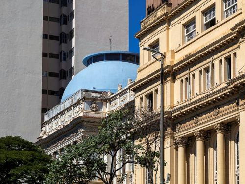 Fachada de edifício antigo bege com colunas e janelas alinhadas. Ao lado, aparece parte de outra construção ornamentada com cúpula azul. Árvores e um poste de iluminação estão em primeiro plano, e prédios altos surgem ao fundo.