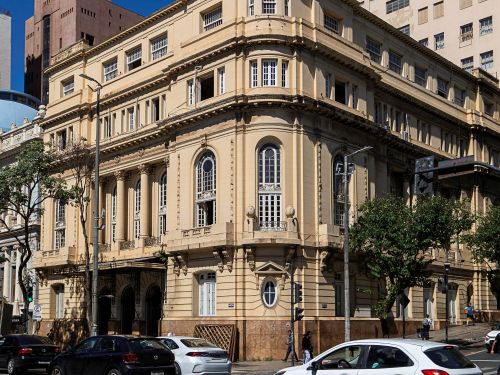 Edifício antigo de fachada bege com janelas altas e detalhes ornamentais ocupa a esquina de uma avenida movimentada. Há árvores na calçada e vários carros circulando na rua em frente. Ao fundo, aparecem prédios altos e o céu azul.